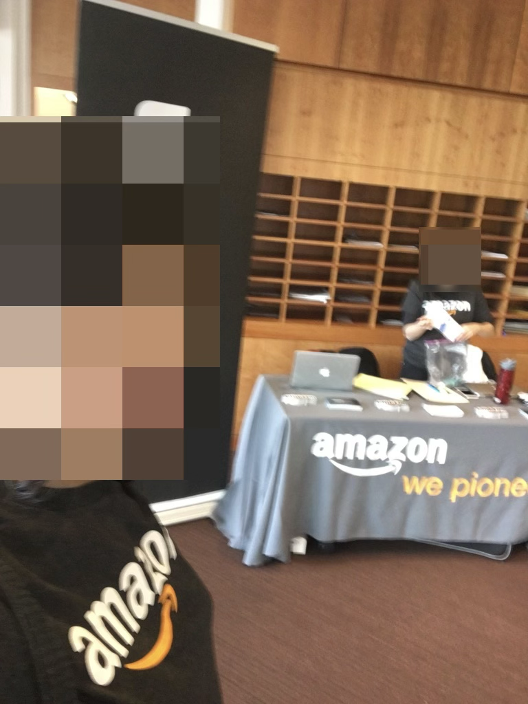 A photo of a woman wearing an Amazon t-shirt next to an Amazon table and an Amazon banner. Faces are blurred out.